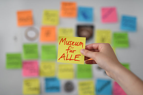 Eine Wand mit bunten Post-it-Zetteln, in der Mitte einen Hand, die einen gelben Zettel mit der Aufschrift Museum für ALLE hält