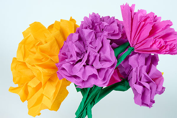 Alt-Text: Bunte Papierblumen aus Krepppapier in Gelb, Rosa und Lila vor hellem Hintergrund. Die floralen Bastelarbeiten erinnern an dekorative Blüten und greifen spielerisch Themen von Kultur und Gestaltung im Badischen Landesmuseum auf.