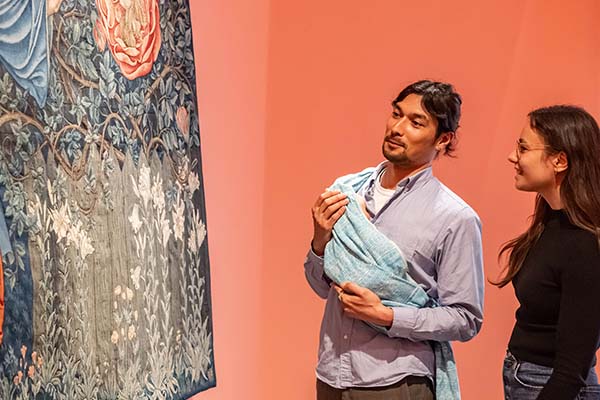 Zwei Besucherinnen und Besucher betrachten in der Ausstellung Bloom up! Die Sprache der Blumen einen großformatigen Wandteppich mit floralen Motiven. Eine Person hält ein Baby in einem Tragetuch. Das Vermittlungsangebot des Badischen Landesmuseums verbindet Kultur, Kunst und gemeinsames Erleben für Erwachsene mit Babys.