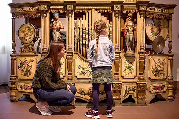 Kind und erwachsene Begleitperson betrachten einen historischen Musikautomaten mit Figuren, Orgelpfeifen und bemalten Dekorelementen im Deutschen Musikautomatenmuseum in Bruchsal. Das reich verzierte Objekt veranschaulicht technische und künstlerische Aspekte vergangener Kultur und ist Teil der Sammlung des Badischen Landesmuseums.