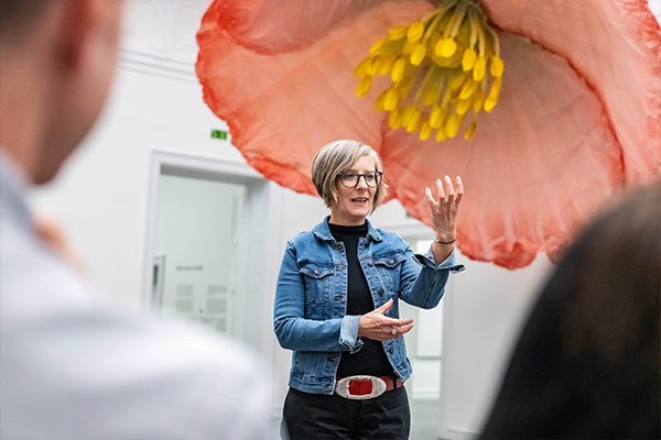 Eine Kulturvermittlerin spricht bei einer Führung des Badischen Landesmuseums vor einer überdimensionalen Blüteninstallation in der Ausstellung Bloom up! Die Sprache der Blumen und gestikuliert vor den Teilnehmenden.