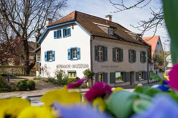 Schrägansicht des Keramikmuseums Staufen im Breisgau mit blauen Fensterläden, umgeben von Bäumen und Blumen im Vordergrund. Das Gebäude steht für keramische Tradition und Kultur und ist Teil des Badischen Landesmuseums.