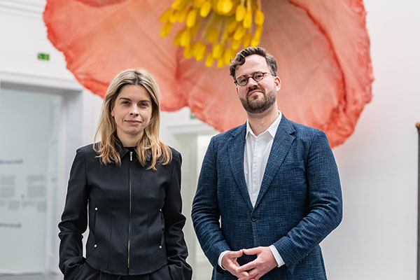 Zwei Kurator:innen des Badischen Landesmuseums stehen vor einer großen Blüteninstallation in der Ausstellung Bloom up! Die Sprache der Blumen und repräsentieren die Zusammenarbeit von Kunst und Kultur.