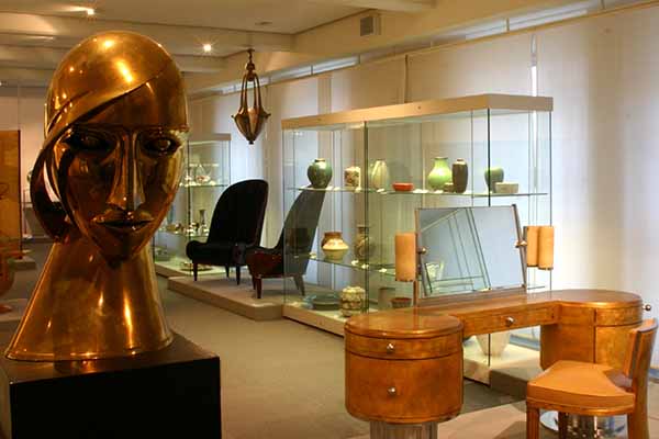 Ausstellungsraum im Museum beim Markt in Karlsruhe mit Art-Déco-Möbeln und Designobjekten, darunter eine große goldfarbene Kopfskulptur sowie Keramik in Vitrinen. Die Präsentation zeigt vielfältige Aspekte von Gestaltung und Kultur im Badischen Landesmuseum.