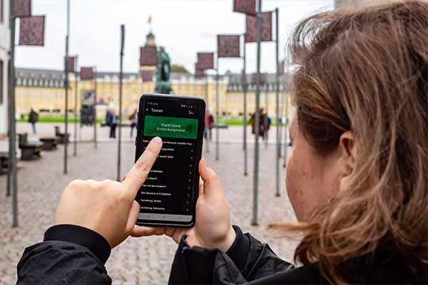 Person nutzt die Museumsapp „PING! Orte der Demokratiegeschichte“ auf dem Smartphone vor dem Karlsruher Schloss. Die Anwendung vermittelt historische Inhalte und verbindet digitale Angebote mit Orten der Kultur des Badischen Landesmuseums.