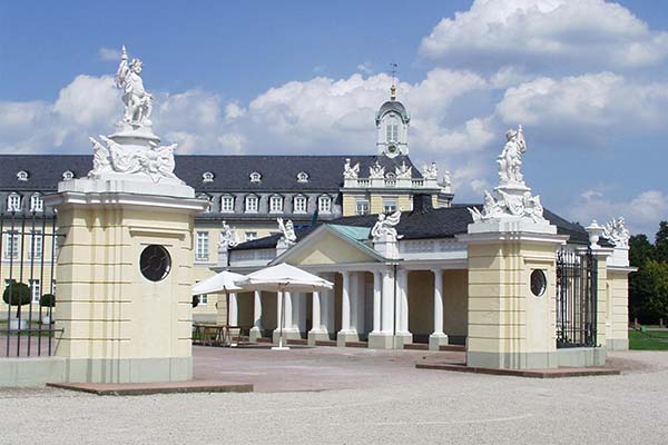 Barocke Schlossanlage mit repräsentativem Torbau, Säulenhalle und reich geschmückten Figuren auf den Torpfeilern unter blauem Himmel. Architekturensemble aus Karlsruhe als Teil der regionalen Geschichte und Kultur, dokumentiert durch das Badische Landesmuseum.
