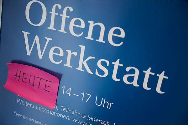 Blaues Schild mit der Aufschrift „Offene Werkstatt“ und einem pinken Zettel mit dem handschriftlichen Hinweis „Heute“. Die Ankündigung verweist auf ein kreatives Kulturangebot des Badischen Landesmuseums.
