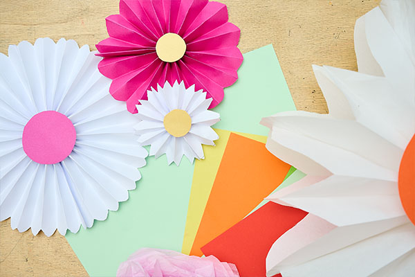 Gefaltete Papierblumen in Weiß, Pink und Rosa liegen neben bunten Papierbögen auf einem Tisch. Die kreativen Bastelarbeiten verbinden florales Gestalten und Kulturvermittlung im Workshopprogramm des Badischen Landesmuseums.