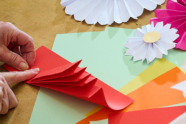 Hände falten rotes Papier zu einer dekorativen Papierform, daneben liegen weitere Papierblumen und bunte Bastelbögen. Das kreative Workshopangebot verbindet florales Gestalten und Kulturvermittlung im Badischen Landesmuseum.