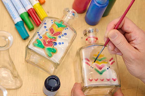 Hand bemalt kleine Glasflaschen mit bunten floralen Mustern, daneben liegen Farben und Stifte auf einem Tisch. Das kreative Workshopangebot verbindet Gestaltung, Handarbeit und florale Kultur im Badischen Landesmuseum.