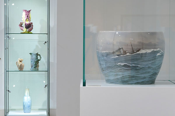 Vitrine mit keramischen Objekten aus der Ausstellung „Jugendstil International“ im Keramikmuseum Staufen, darunter eine große Vase mit maritimer Darstellung eines Schiffes in bewegter See sowie kleinere Gefäße und plastische Arbeiten. Präsentation von Keramik und Gestaltung als Teil der Kultur im Badischen Landesmuseum.