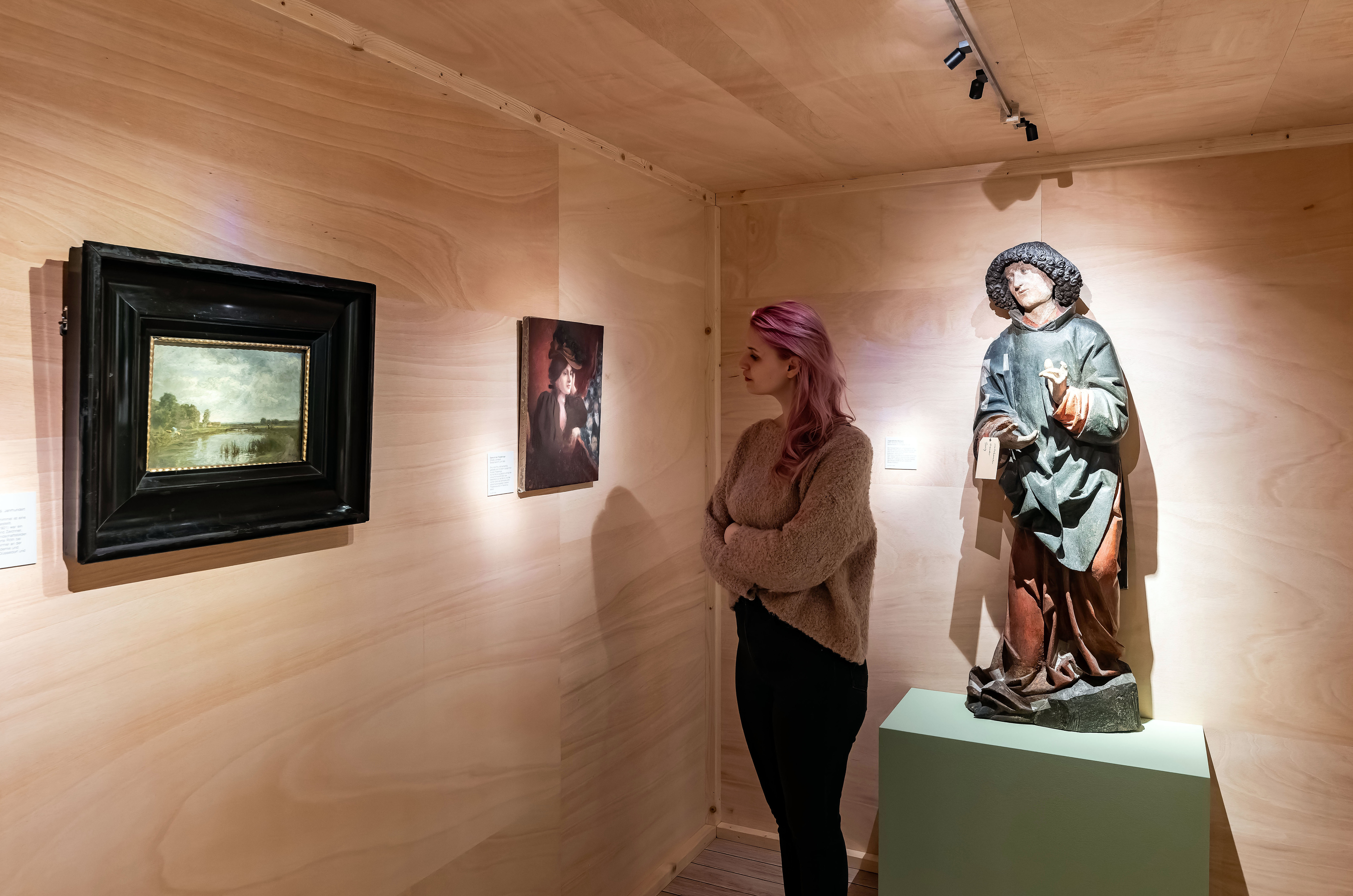 Besucherin betrachtet ein kleines Gemälde, hinter ihr steht die bemalte Holzskulptur eines Heiligen auf einem Sockel