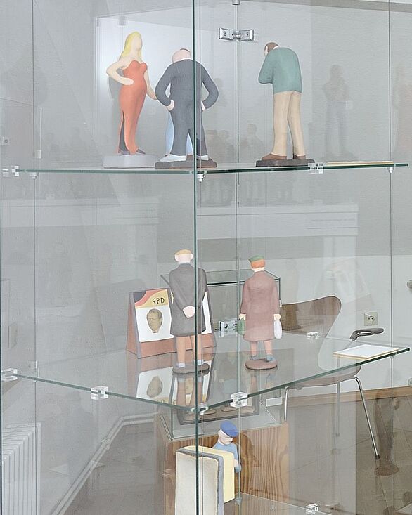 Eine Glasvitrine im Keramikmuseum Staufen zeigt mehrere stilisierte Figuren von Elisabeth Winter-Bonn auf unterschiedlichen Ebenen. Die farbig gestalteten Keramikskulpturen stellen Menschen in alltäglichen Körperhaltungen dar, darunter eine Frau im roten Kleid und verschiedene männliche Figuren. Das Badische Landesmuseum präsentiert diese Werke im Kontext zeitgenössischer Keramikkultur.