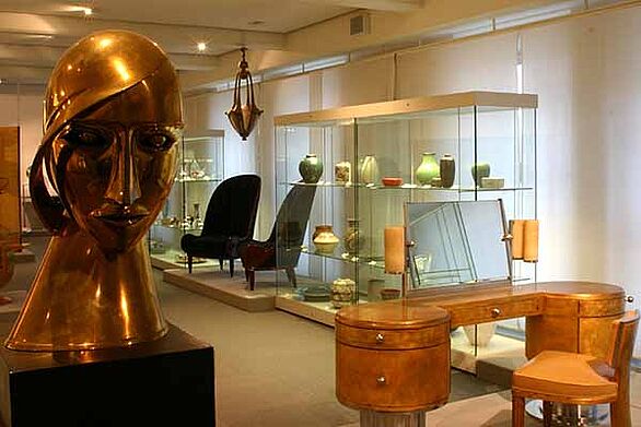 Ausstellungsraum im Museum beim Markt in Karlsruhe mit Art-Déco-Möbeln und Designobjekten, darunter eine große goldfarbene Kopfskulptur sowie Keramik in Vitrinen. Die Präsentation zeigt vielfältige Aspekte von Gestaltung und Kultur im Badischen Landesmuseum.