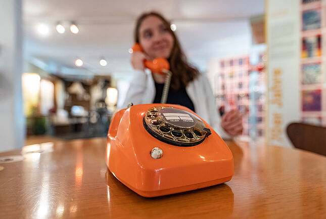 Eine Person hält sich den Hörer eines orangenen Wählschreibentelefons an das Ohr.
