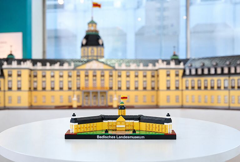 Mini Schloss Karlsruhe aus Klemmbausteinen auf einem weißen Tisch: Das Modell des Badischen Landesmuseums zeigt die gelbe Schlossfassade, schwarze Dächer und den Schlossturm mit badischer Flagge. Im Hintergrund ist ein größeres Modell des Karlsruher Schlosses zu sehen. Das Mini Schloss verbindet Architektur, Geschichte und Kultur im Kleinformat.