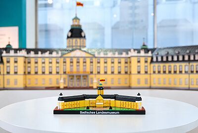 Mini Schloss Karlsruhe aus Klemmbausteinen auf einem weißen Tisch: Das Modell des Badischen Landesmuseums zeigt die gelbe Schlossfassade, schwarze Dächer und den Schlossturm mit badischer Flagge. Im Hintergrund ist ein größeres Modell des Karlsruher Schlosses zu sehen. Das Mini Schloss verbindet Architektur, Geschichte und Kultur im Kleinformat.