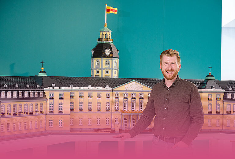 Mann steht vor dem Klötzle-Schloss, einem detailreichen Modell des Karlsruher Schlosses aus Klemmbausteinen. Im Hintergrund ist der Schlossturm mit badischer Flagge zu sehen. Das Modellprojekt des Badischen Landesmuseums verbindet Architektur, Kultur und Miniaturbau.