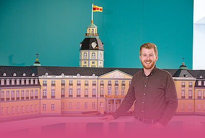 Mann steht vor dem Klötzle-Schloss, einem detailreichen Modell des Karlsruher Schlosses aus Klemmbausteinen. Im Hintergrund ist der Schlossturm mit badischer Flagge zu sehen. Das Modellprojekt des Badischen Landesmuseums verbindet Architektur, Kultur und Miniaturbau.