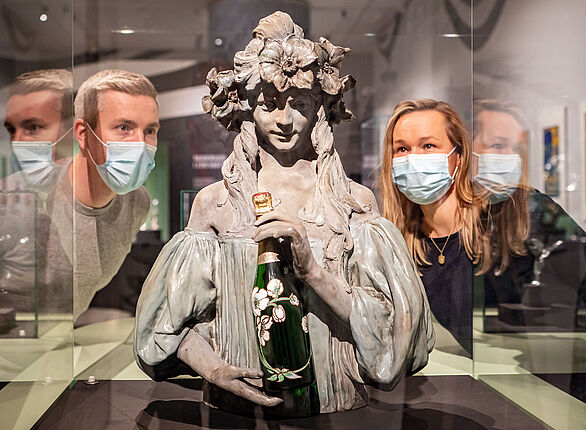 Zwei Personen betrachten die Skulptur einer jungen Frau, die eine Champagnerflasche in den Händen hält.