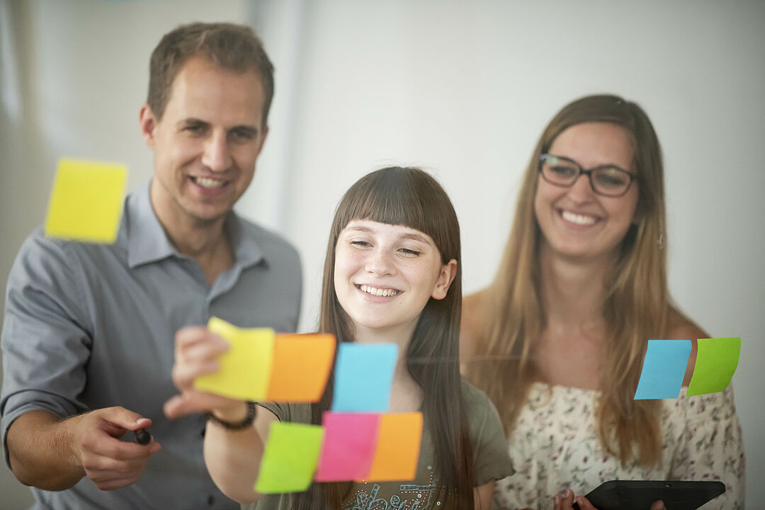 Ein Mann und zwei Frauen in einer Workshop-Situation: sie kleben bunte Post-its mit Ideen an eine Fenster