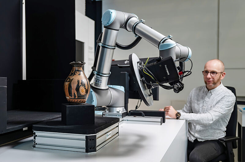 eine antike Vase wird von einem 3D-Scanner gescannt, dahinter sitzt ein Mann in einem weißen Kittel