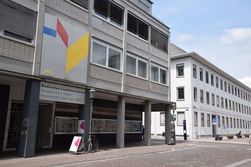 Außenansicht des Museum beim Markt in Karlsruhe mit moderner Fassade und Eingangsbereich unter Arkaden. Über dem Eingang hängt ein Schild mit der Aufschrift Badisches Landesmuseum Museum beim Markt Angewandte Kunst seit 1900. Vor dem Gebäude stehen ein Fahrrad und ein Aufsteller. Das Badische Landesmuseum Karlsruhe präsentiert hier Kultur, Design und vielfältige Veranstaltungen im historischen Stadtkern.