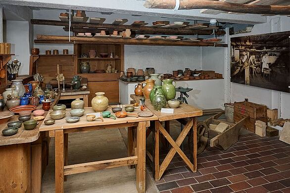 Historische Töpferwerkstatt im Keramikmuseum Staufen des Badischen Landesmuseums. Auf massiven Holztischen stehen zahlreiche glasierte Krüge, Vasen, Schalen und Teller. Werkzeuge, Holzregale, Arbeitsmaterialien und eine historische Fotografie ergänzen die Einrichtung. Die Ausstellung vermittelt Einblicke in das traditionelle Töpferhandwerk und die regionale Keramikkultur.