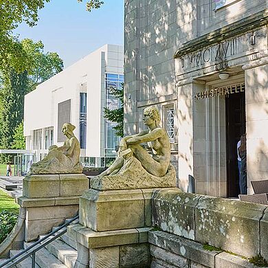 Eingangsbereich der Staatlichen Kunsthalle Baden-Baden mit zwei historischen Sitzfiguren aus Stein vor der Fassade. Im Hintergrund ist der moderne Erweiterungsbau des Museums zu sehen. Die Kunsthalle dient dem Badischen Landesmuseum als Interimsquartier und verbindet Architektur, Kunst und Kulturgeschichte.