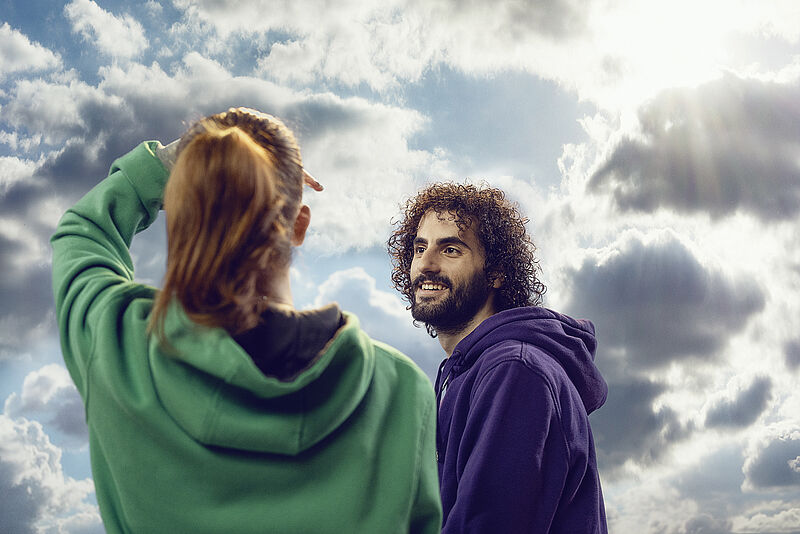 Die Rückenansicht einer Frau, die in den Himmel und die Sonne blickt und ein fröhlicher Mann mit schwarzen Locken der sich ihr zuwendet