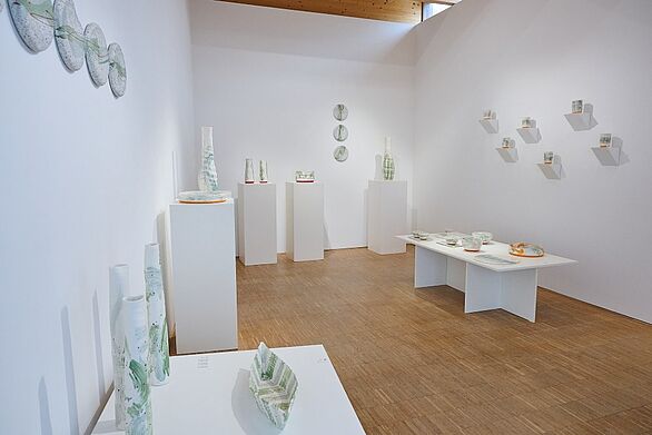Blick in eine Studioausstellung des Keramikmuseums Staufen, einem Standort des Badischen Landesmuseums. In einem hellen Ausstellungsraum mit Holzboden stehen filigrane Keramikvasen, Schalen und Teller mit überwiegend weißen und grünen Glasuren auf Podesten, Wandregalen und einem niedrigen Tisch. Die Präsentation zeigt die gestalterische Vielfalt zeitgenössischer Keramikkultur.