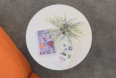 Auf einem runden weißen Tisch im Museum beim Markt liegen die Bücher „Schwebende Lasten“ von Annett Gröschner und „Wachs“ von Christine Wunnicke neben einer Pflanze. Die LeseZeit ist das neue Buchclub-Format des Badischen Landesmuseums und lädt zum gemeinsamen Austausch über Literatur und Kultur ein.
