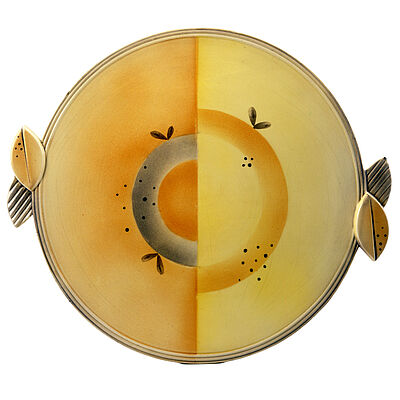 Keramische Tortenplatte mit seitlichen Griffen und geometrischem Dekor in Gelb, Orange und Grau. Die kreisförmige Gestaltung ist in zwei Farbflächen geteilt und mit Punkten sowie stilisierten Blättern verziert. Objekt aus der Sammlung des Badischen Landesmuseums und ein Beispiel für Keramikdesign und Kultur des frühen 20. Jahrhunderts.