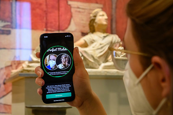 Eine Nutzerin mit ihrem Smartphone, auf dem Ping! Die Museumsapp geöffnet ist. Der Screen zeigt ein Match mit einer liegenden Etruskerin - dem Objekt, das die im Hintergrund zu sehen ist.