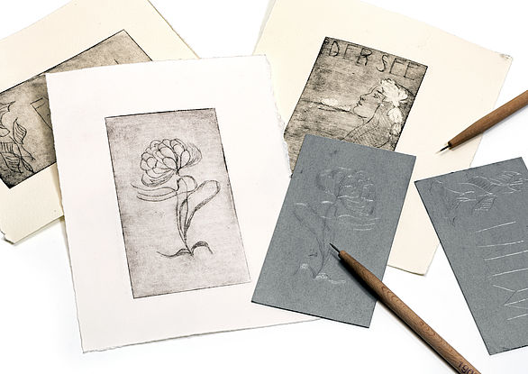 Radierungen zum Thema Jugendstil: Drei Karten, zwei Druckplatten und zwei Bleistifte liegen übereinander. Im Vordergrund zu sehen ist eine fertige Radierung auf einer Karte, die eine Blume zeigt.