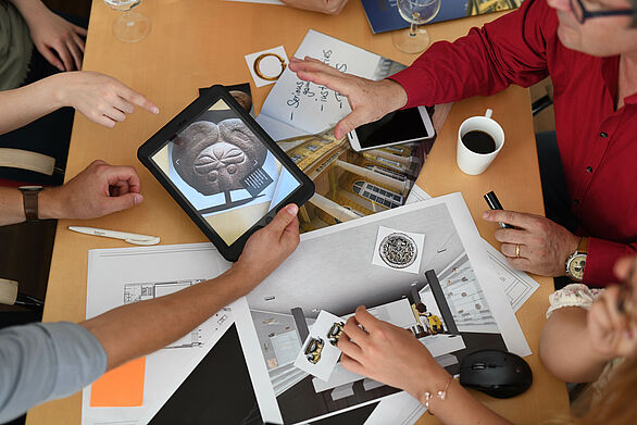 Workshop: Ein Tisch, mehrere Hände, Kaffeetassen, vielen Arbeitsmaterialien. Papier und einem Tablet. 