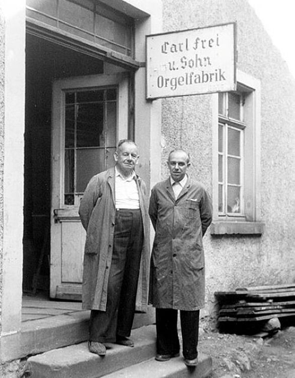 Historische Schwarz-Weiß-Fotografie zweier Männer vor der Orgelfabrik Carl Frei und Sohn in Waldkirch. Das Bild zur Ausstellung „Zuhause – Unterwegs“ im Deutschen Musikautomaten-Museum des Badischen Landesmuseums verweist auf die Kultur und Tradition des Orgelbaus sowie die internationale Geschichte mechanischer Musik.