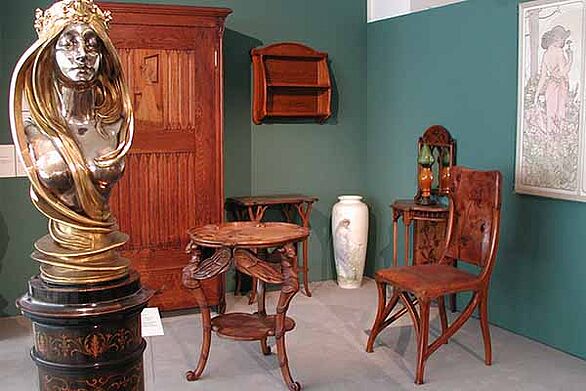 Ausstellungsraum im Museum beim Markt in Karlsruhe mit Jugendstil-Möbeln, darunter ein kunstvoller Holzstuhl, ein kleiner Tisch und ein Schrank sowie eine metallische Büste. Die Präsentation zeigt Gestaltung und Kultur des Jugendstils im Badischen Landesmuseum.