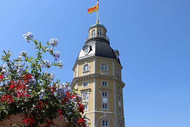 Der Schlossturm des Badischen Landesmuseums, auf dem die badische Flagge vor blauem Himmel weht. Im Vordergrund sind Blumen zu sehen.