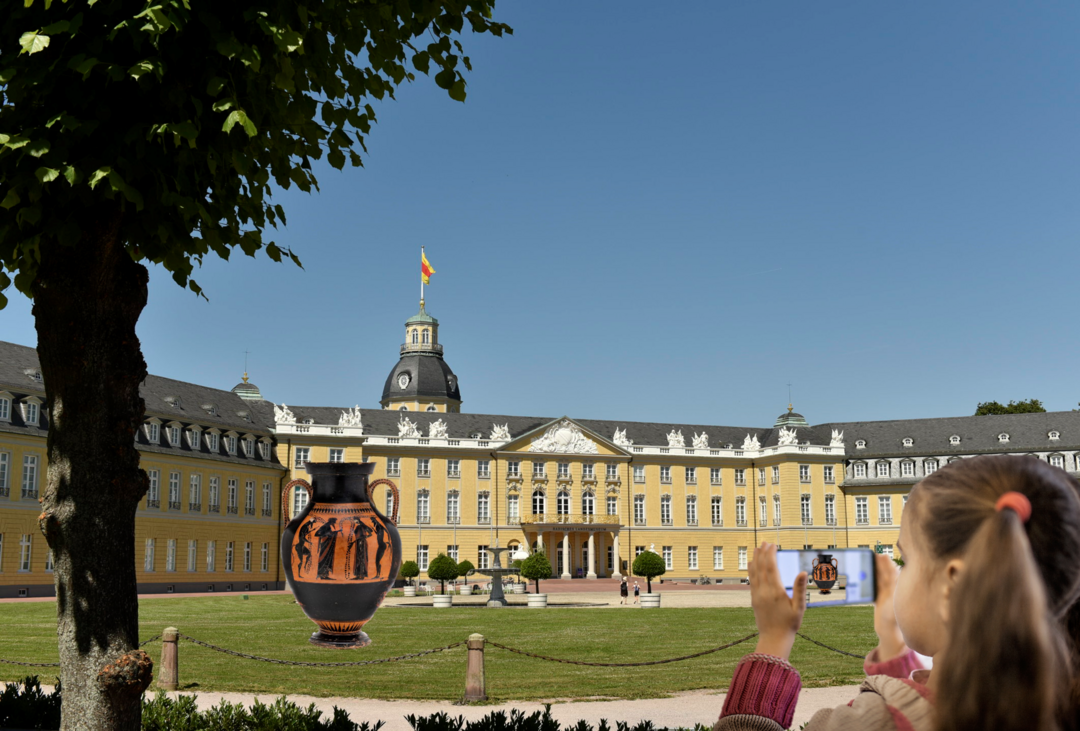 Ein Kind betrachtet auf einem Smartphone eine Vase, die per Augumented Reality vor der Karlsruher Schloss platziert ist.