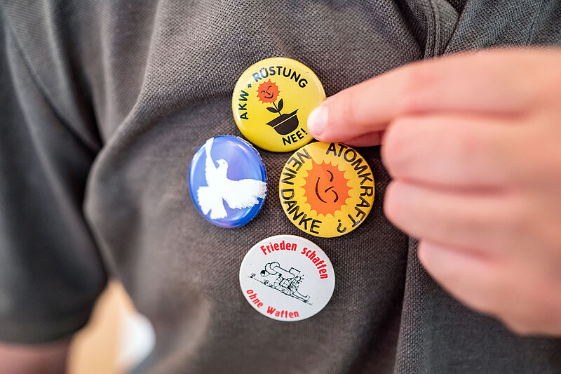 Vier bunte Ansteckbuttons, die an einem grauen Hemd befestigt sind.