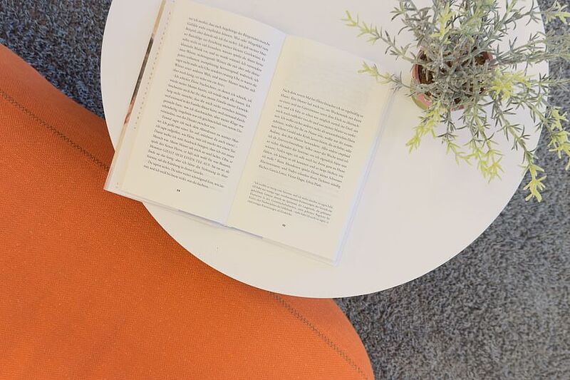 Ein aufgeschlagenes Buch liegt auf einem runden weißen Tisch neben einer grünen Topfpflanze und einem orangefarbenen Sitzpolster. Das Motiv steht für LeseZeit, den Buchclub im Museum beim Markt des Badischen Landesmuseums in Karlsruhe. Die Veranstaltung verbindet Literatur, gemeinsames Lesen und Gespräche über Kultur in gemütlicher Atmosphäre.