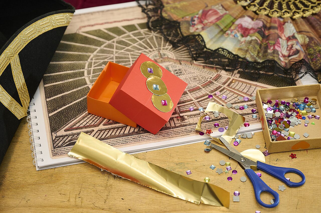 Ein mit Goldfolie und Glitzersteinen gestaltetes Schatzkästschen liegt neben einem Fächer, daneben eine Schatulle mit vielen weiteren Glitzersteinen, Goldfolie und einer Schere