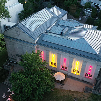 Luftaufnahme der Staatlichen Kunsthalle Baden-Baden am Abend. Das klassizistische Gebäude ist von Bäumen umgeben, die Fensterfront leuchtet in pinken und gelben Farben. Die Kunsthalle dient dem Badischen Landesmuseum derzeit als Interimsquartier für Kultur und Ausstellungen.