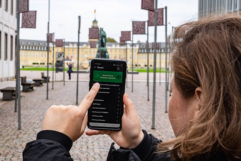 Eine Frau bedient ihr Smartphone, auf dem die App Ping läuft, im Hintergrund ist das Karlsruher Schloss