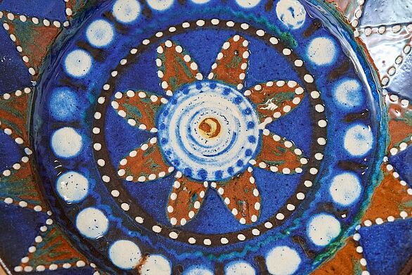Detailaufnahme eines Keramikmusters von Josef Maier im Keramikmuseum Staufen. Eine sternförmige Blüte mit rotbraunen Blättern, weißen Punkten und konzentrischen Kreisen schmückt die intensiv blau glasierte Oberfläche. Das Badische Landesmuseum dokumentiert mit diesem kunstvollen Dekor die Vielfalt keramischer Gestaltung und Kultur.