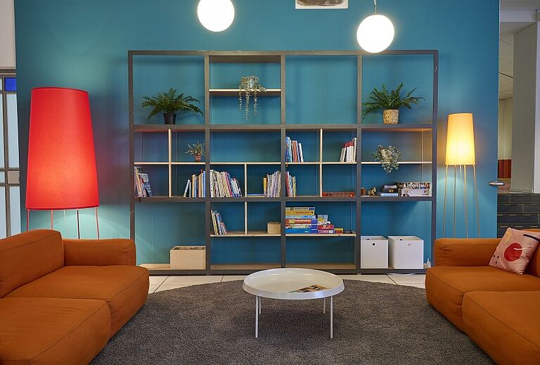 Der frei zugängliche Open Space im Museum beim Markt des Badischen Landesmuseums in Karlsruhe ist ein Ort für Kulturveranstaltungen. Zwei orangefarbene Sofas stehen vor einer türkisfarbenen Wand mit einem Regal voller Bücher, Spiele und Pflanzen.