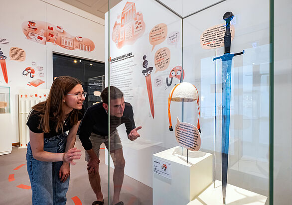 Zwei Personen betrachten eine Vitrine, in der ein Schwert und ein Fahrradhelm ausgestellt sind